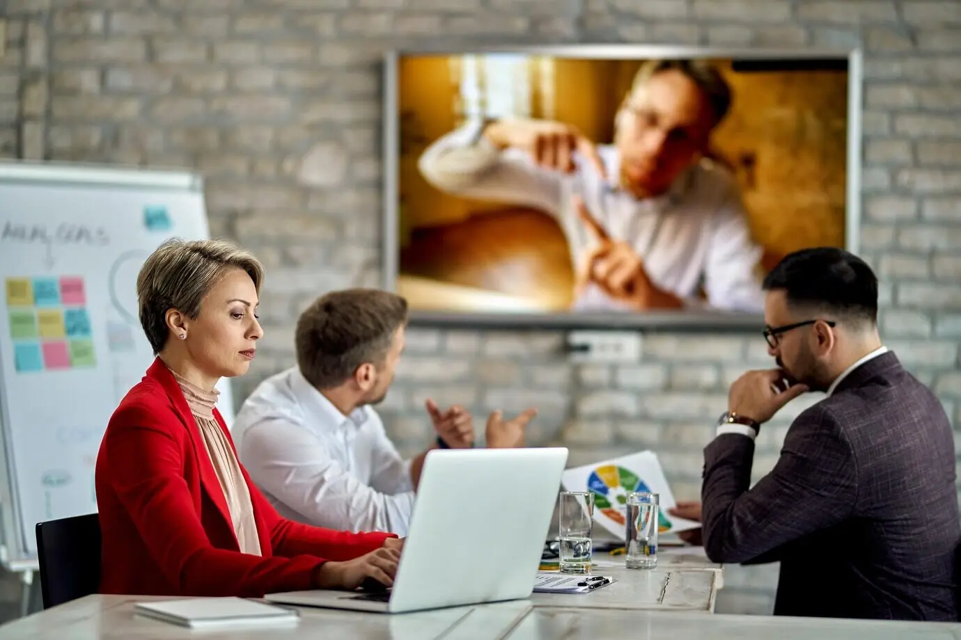 Деловая женщина работает за компьютером во время встречи в офисе; один из её коллег присоединяется к встрече по видеосвязи.